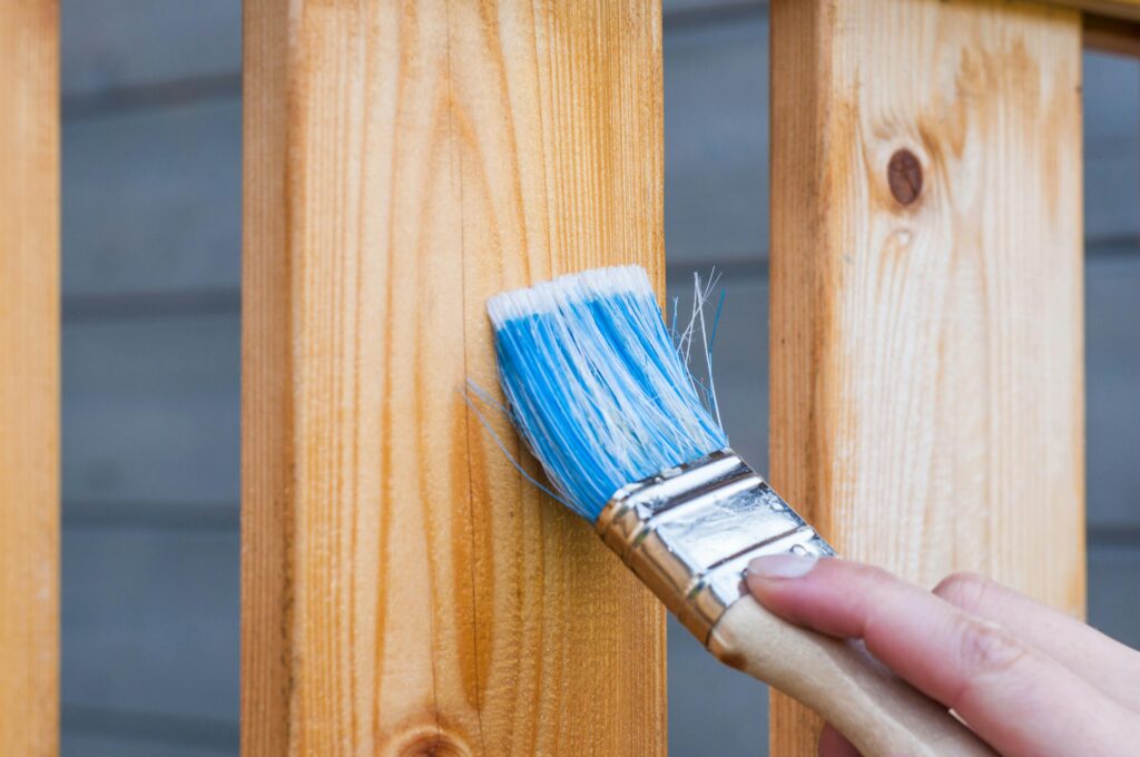 A hand painting a wooden fence plank with a blue paintbrush outdoors. Perfect for DIY and renovation themes. service in Conroe Texas and Montgomery County